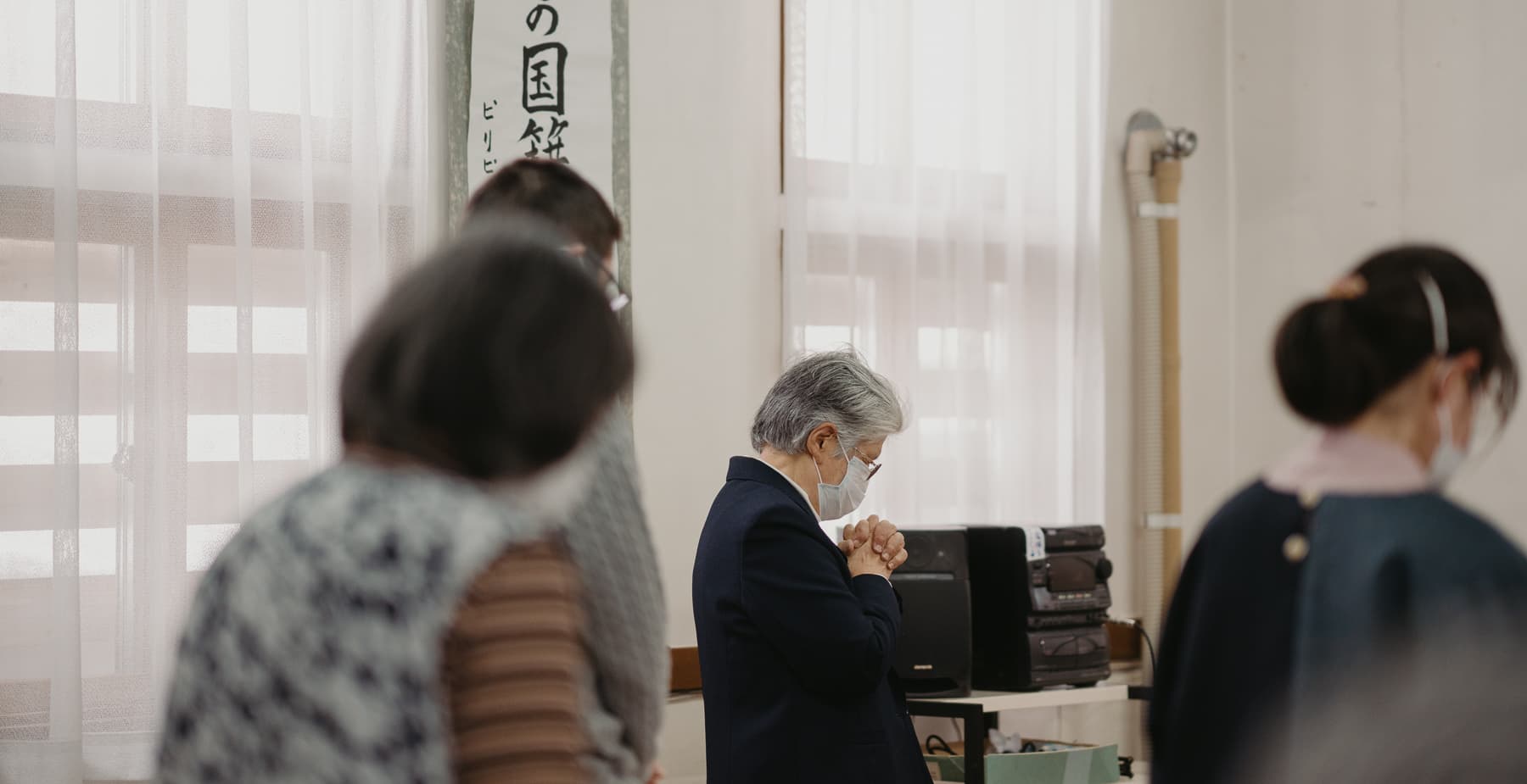 An elderly woman at a church service, standing with her hands folded in prayer.